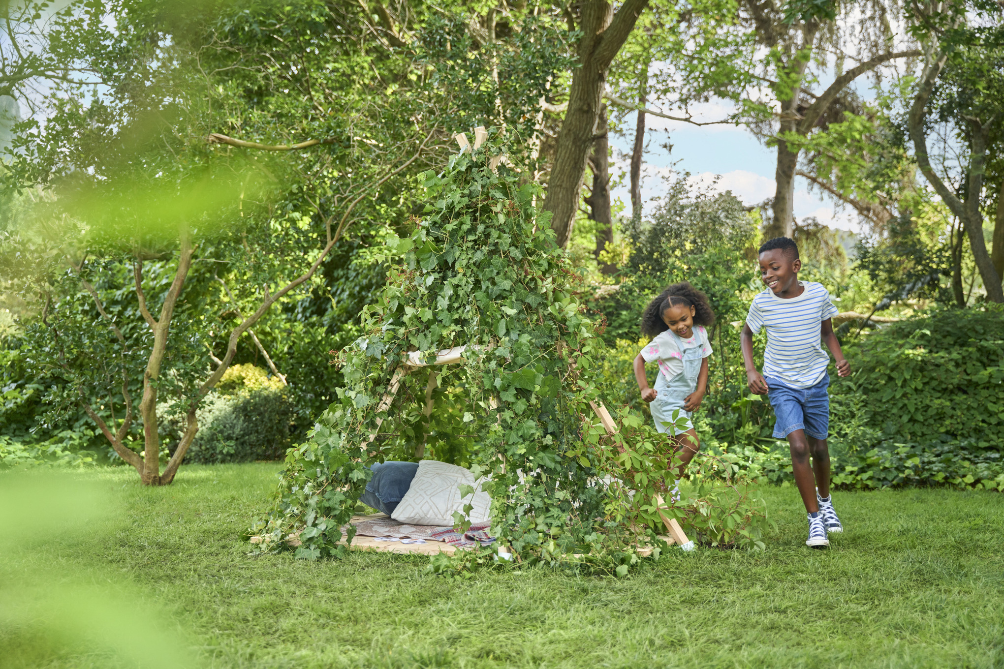 Mädchen und Junge springen um ihr Kinderzelt herum, das mit Kletterpflanzen bedeckt ist, die Decke wurde entfernt 