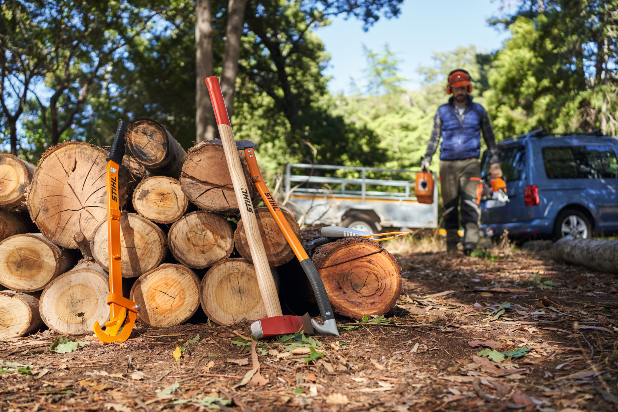 Mann in Schutzausrüstung und mit Geräten in der Hand läuft auf einen Brennholzstapel zu, an dem eine STIHL Forstaxt und weitere Geräte zum Holzspalten lehnen
