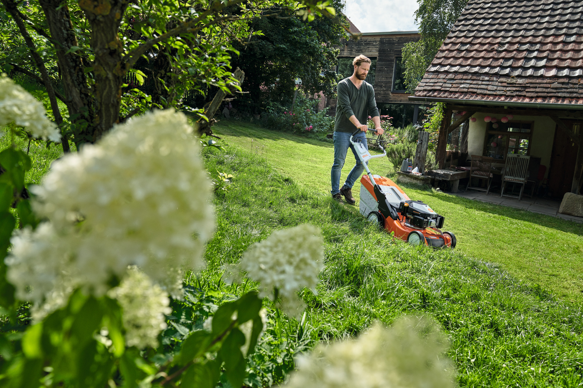 Mann steht mit STIHL Akku Heckenschere im Garten und ein Cashback 100€ Logo ist zu sehen.