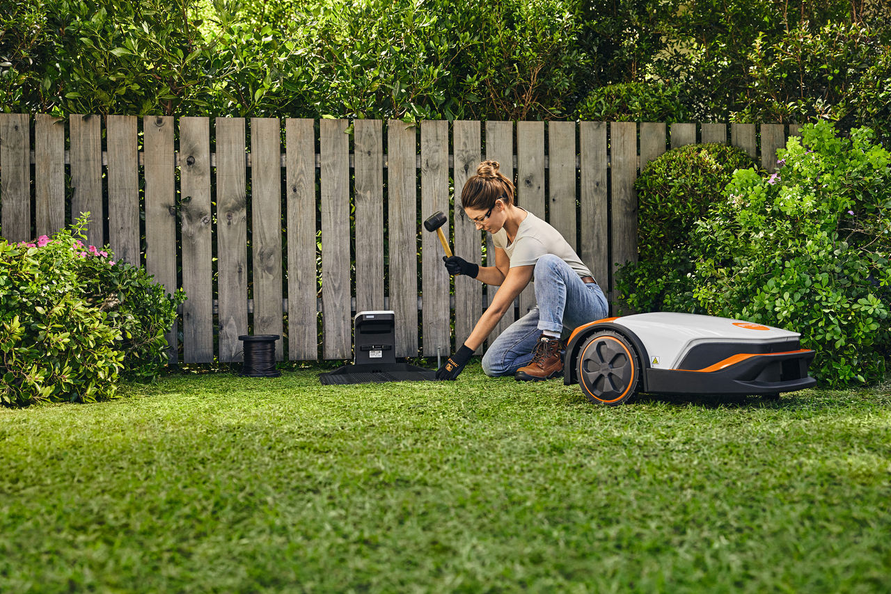 Eine Frau auf einem Gartengrundstück baut die iMOW® Dockingstation auf, dafür hat sie einen Hammer in der Hand